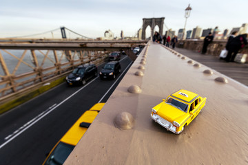 Model of a classic yellow taxi on a steel beam on Brooklyn Bridge, with a real taxi passing on the...