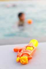 Water gun isolated on the beach chair with abstract blur of a boy playing his water gun in the swimming pool on the background. Shallow depth of field. Concept of summer and Thai water festival.