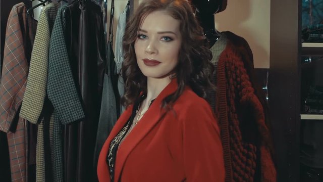 A young brunette woman tries on a red jacket, straightens it and turns looking at the camera next to the racks of ready-made female clothes in the store.