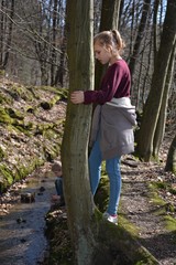 Mädchen am Baum schaut in den Bach, kleiner Junge spielt hinten am Wasser