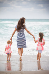 Happy family enjoying vacations by the sea. Mother with two little girl walking on the sandy beach. Back view on young woman with two siblings children. outdoors.