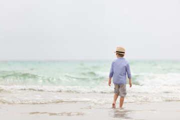 Adorable kid boy in straw hat and sun glasses walking on ocean beach and playing with waves. Vacations by the sea. Outdoor.