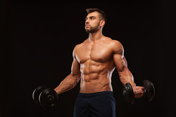 Fototapeta na wymiar Handsome athletic man in gym is pumping up muscles with dumbbells in a gym. Fitness muscular body isolated on dark background.
