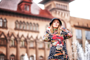 A beautiful girl in a bright multi-colored coat in rainy weather against a bright brick old castle or a house. Fountain in the background. Saratov, Russia.