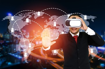 Young asian businessman in black suit wearing VR virtual reality headset, business technology concept for future on world map with social media connection icon on blurred night city background.