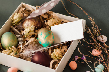Handmade easter gift box with painted eggs, feathers and flowers on grey background. Empty white greeting card with free space for text