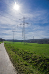 Strommast mit Sonne vor strahlend blauem Himmel, vertikal, im Hintergrund Windkrafträder