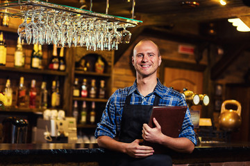 Barman at work in pub,Portrait of cheerful barman worker standing,Waiter giving menus,A pub.Bar.Restaurant.Classic.Evening.European restaurant.European bar.American restaurant.American bar.