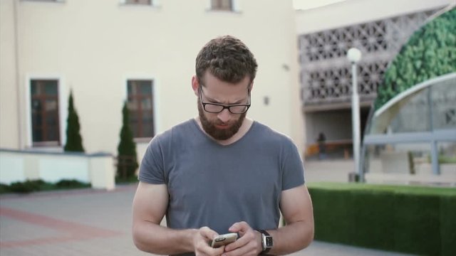 Man with beard and glasses walking in city centre and using smarphone. Guy texting with someone, smiling. Slow motion.