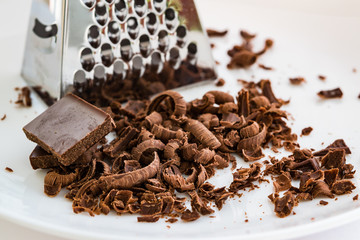 Pieces of grated dark chocolate next to a metal grater on a white plate close up
