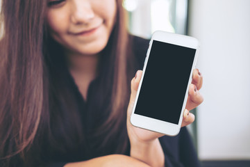 Mockup image of a beautiful woman with smiley face holding and showing white mobile phone with blank black screen  in modern cafe