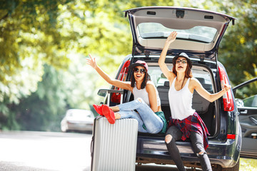 Two young women with suitcases on car trip.They are sitting in car back and resting after long ride and making fun.Coffee break.
