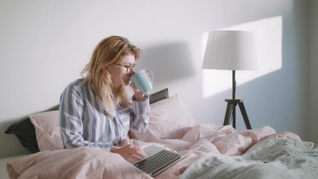 Young female freelancer is sipping on coffee while sitting in the bed and is softly smiling while scrolling through her news feed and checking emails