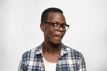 Fototapeta premium Studio shot of stylish African male in glasses staring at camera, having crazy look. Funny young dark-skinned hipster posing at blank wall with copy space for your text or advertising content