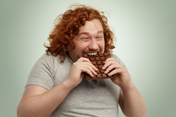 Plump funny redhead young Caucasian male with curly hair biting large of chocolate with pleased...