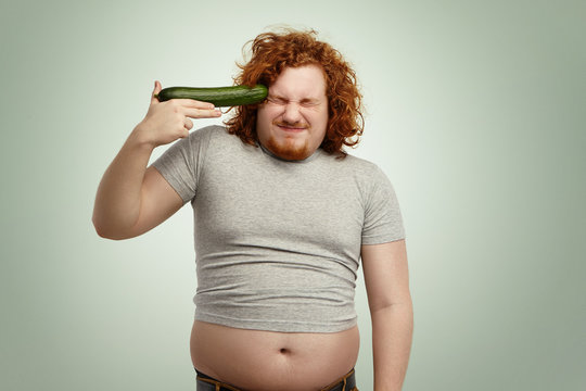 Desperate crazy obese young Caucasian man with ginger hair wearing undersize t-shirt holding cucumber at his temple like gun, ready to shoot himself, keeping eyes closed, pissed off with diet