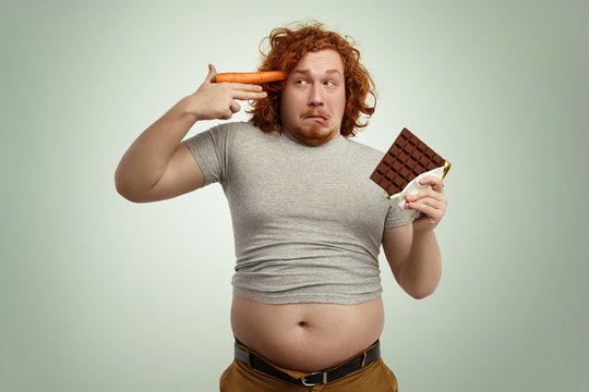 These vegetables are going to kill me. Funny overweight man holding bar of chocolate in one hand and carrot at temple like gun, about to shoot himself, looking with scared and frightened expression