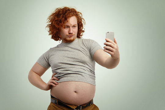 Funny overweight man with fat tummy hanging out of grey t-shirt, standing at studio wall, keeping hand on waist while posing for picture, taking selfie on mobile phone, trying to look seductive