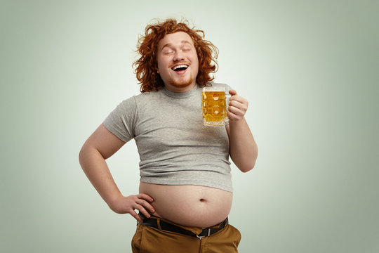 Happy joyful young overweight man with curly red head closing eyes in enjoyment, anticipating first gulp of cold frothy beer, holding glass of beverage in hands, his belly hanging out of pants