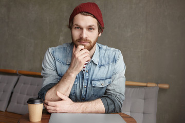 People and lifestyle concept. Handsome confident young Caucasian bearded freelancer in jeans jacket and hat enjoying coffee break while relaxing after having finished work on laptop computer