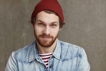 Close up shot of charismatic fashionable young European male with thick beard wearing trendy hat...