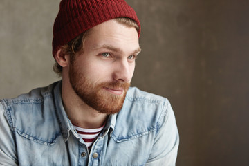 Indoor portrait of stylish good-looking young bearded hipster in trendy clothes with joyful mysterious smile, sitting at blank concrete copy space wall for your content, thinking about his girlfriend