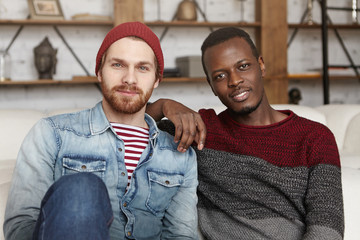 Happy homosexual interracial couple having rest indoors. African American male resting elbow on shoulder of his handsome stylish partner in trendy clothes, both looking at camera with joyful smiles
