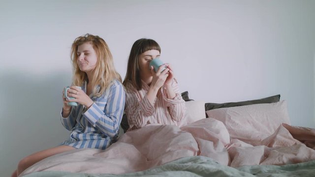 A blonde young woman is bringing her beautiful brunette partner coffee or tea in bed in bright and full of light room, they talk and laugh playfully