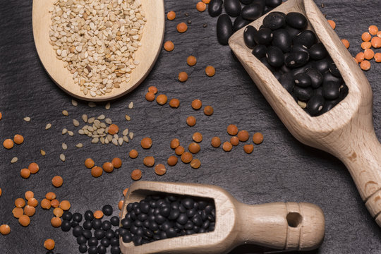 Sesame seeds, red and black lentils and black beans with wooden spoons on black background