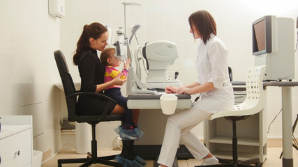Mother and daughter in the ophthalmologist's room- optometrist in clinic checking little child's...