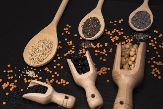 Sesame, chia and poppy seeds, red and black lentils, chickpeas and black beans with wooden spoons on black background