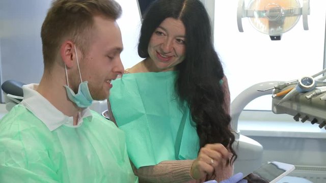 Senior brunette woman looking at the dentist's tablet. Mature caucasian lady pointing her finger on the screen. Young blond dental hygienist answering his client's questions