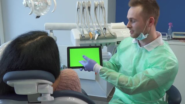 Handsome young male dentist pointing his hand on screen with green chroma key. Attractive caucasian dental hygienist holding tablet in his hand. Female client sitting backwards on the dental chair