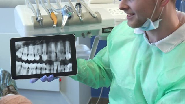 Attractive caucasian dentist in medical uniform moving x-ray image on his tablet. Close up of young blond dental hygienist sitting against background of different dental handpieces. Camera tilting up