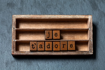 Je t'adore. I love adore you in French translation. Vintage box, wooden cubes phrase written with old style letters. Gray stone textured background. Close-up, up view, soft focus