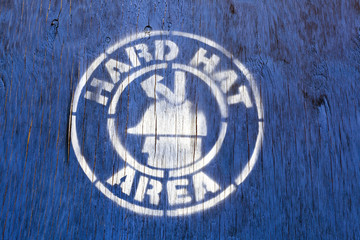 HARD HAT AREA construction site sign stenciled on blue wood panel.