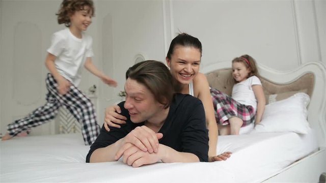 Restless and Active Family Playing Together on a Bed at Home During Weeked. Young Parents with Charming Smiles are Spending Free Time with Their Son and Daughter in the Bedroom in the Morning.