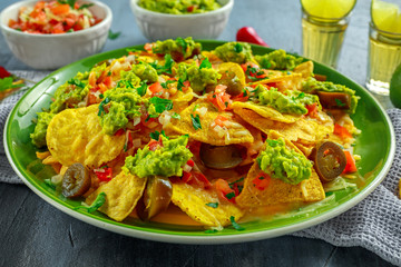 Nachos with cheese, jalapeno peppers, red onion, parsley, tomato, salsa, guacamole sauce and tequila on green plate.