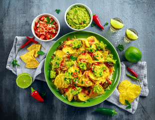 Nachos with cheese, jalapeno peppers, red onion, parsley, tomato, salsa, guacamole sauce and tequila on green plate.