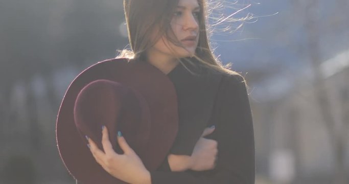 4k. Attractive young girl with a long hair walks in the sun and wind blowing, holding purple hat, autumn coat, stedicam portrait shot