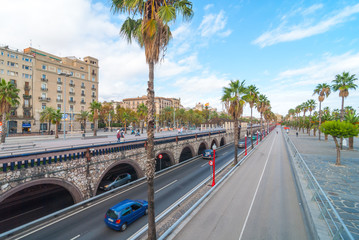 Late afternoon in Seaside Barcelona, Spain, sees traffic in tunnelled streets and pedestrian traffic above as people enjoy walking outdoors along the waterfront in warm weather in November.