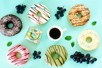 Morning coffee with sweets over light blue background. Assorted donuts (pistachio, caramel, strawberry, chocolate, vanila), grapes and cup of black coffee. 