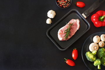 Stylish meat background. Raw pork or beef steak in the pan, bell pepper, cherry tomatoes, broccoli, mushrooms over black board. Copy space.