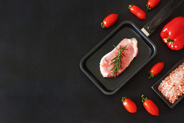 Raw pork steak in the pan with salt, pepper and tarragon. Fresh vegetables (bell pepper, broccoli, cherry tomatoes) over black board. Dinner ingredients. Copy space.