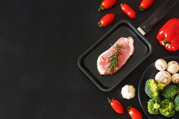 Stylish meat background. Raw pork or beef steak in the pan, bell pepper, cherry tomatoes, broccoli, mushrooms over black board. Copy space.