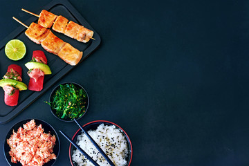 Top view of tuna sushi, grilled salmon on skewers, bowl or rice, crab meat and wakame salad over dark board with a copy space. Gourmet japanese dinner. 