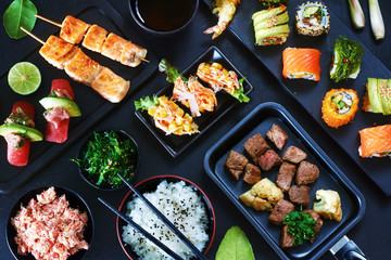 Top view of japanese style meal over dark background. Tuna sushi, assorted sushi rolls with salmon, avocado, crab, slice beef steak, wakame, crab salad and green tea. 