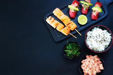 Grilled salmon on skewers, tuna sushi, crab salad, wakame and bowl of rice over dark background with copy space. Top view of stylish japanese meal. 