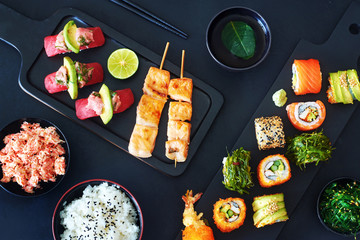 View from above of tuna sushi, different sushi rolls with salmon, shrimp, avocado, wakame,  grilled salmon on skewers, crab meat, bowl of rice and cup of green tea. Dark background. 