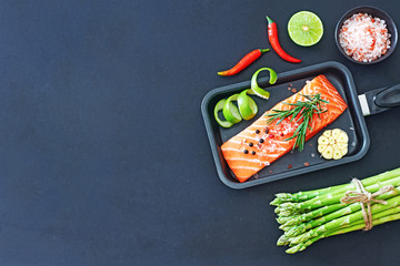 Raw salmon steak and green asparagus with chili, lime, salt and garlic ready to be cooked over dark background. Healthy dinner ingredients concept. Copy space.